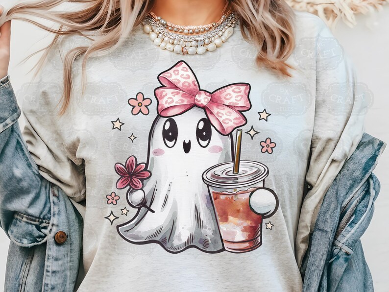 May include: A light gray t-shirt with a cartoon ghost wearing a pink bow and holding a cup of coffee. The ghost has big eyes and a happy expression. The shirt is decorated with small pink flowers and stars.
