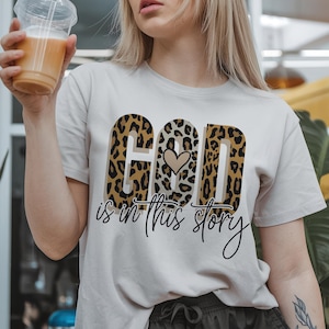 Puede incluir: Una camiseta blanca con un dise&ntilde;o de estampado de leopardo que dice "Dios est&aacute; en esta historia" con un peque&ntilde;o coraz&oacute;n en el medio de la palabra "Dios".