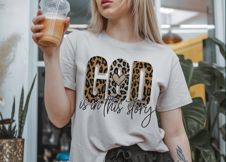 Puede incluir: Una camiseta blanca con un dise&ntilde;o de estampado de leopardo que dice "Dios est&aacute; en esta historia" con un peque&ntilde;o coraz&oacute;n en el medio de la palabra "Dios".