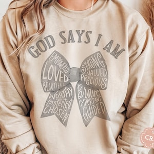 May include: Beige sweatshirt with the text "GOD SAYS I AM" above a bow design. The bow includes words like "LOVED", "STRONG", "BRAVE", and "CHOSEN". The sweatshirt has long sleeves and a relaxed fit.
