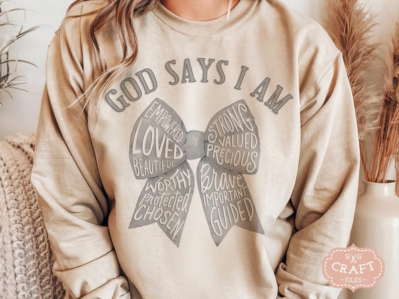 May include: Beige sweatshirt with the text "GOD SAYS I AM" above a bow design. The bow includes words like "LOVED", "STRONG", "BRAVE", and "CHOSEN". The sweatshirt has long sleeves and a relaxed fit.