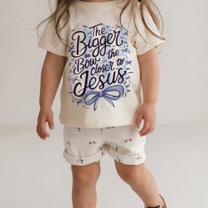 May include: A child's cream-colored t-shirt and shorts set. The t-shirt features the text "The Bigger the Bow Closer to Jesus" in blue script with a blue bow graphic. The shorts are white with a small red and white pattern.