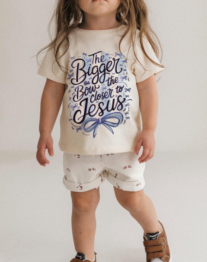 May include: A child's cream-colored t-shirt and shorts set. The t-shirt features the text "The Bigger the Bow Closer to Jesus" in blue script with a blue bow graphic. The shorts are white with a small red and white pattern.
