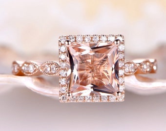 May include: A rose gold engagement ring with a square-shaped morganite center stone surrounded by a halo of smaller diamonds. The band is also set with smaller diamonds in a delicate, vintage-inspired design.