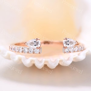 May include: Rose gold ring featuring two pear-shaped, clear gemstones and a band encrusted with small, round, clear gemstones. The ring has an open design, with the pear-shaped stones positioned at the top.