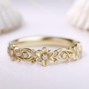 May include: A gold band ring featuring a repeating floral motif with engraved petals and swirling vine details. Small, clear, round-cut stones are set into the center of each flower, creating a delicate, continuous pattern around the entire ring.