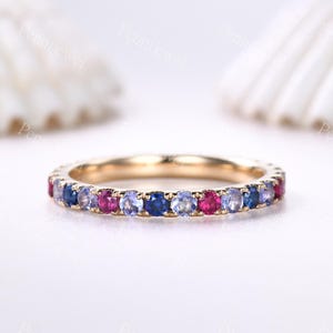 Op de afbeelding: Een gouden bandring met een doorlopende rij kleurrijke edelstenen. De edelstenen wisselen af tussen tinten blauw, paars en roze. De ring is geplaatst tegen een witte achtergrond.