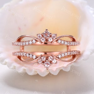 K&ouml;nnte beinhalten: Ros&eacute;goldener Ringverst&auml;rker mit einem zarten, kronenartigen Design, das mit einer Gruppe kleiner, klarer Steine besetzt ist. Der Verst&auml;rker ist mit einer Reihe kleinerer Steine entlang der Band besetzt.
