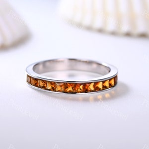 May include: A thin silver band with a row of small, square-shaped, orange gemstones.