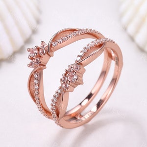 K&ouml;nnte beinhalten: Ros&eacute;goldring mit einem Doppelband-Design. Das Band ist mit kleinen, klaren Steinen besetzt und verf&uuml;gt &uuml;ber ein gr&ouml;&szlig;eres, blumenf&ouml;rmiges Design mit rosafarbenen Steinen.