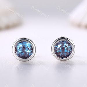 May include: A pair of round stud earrings with a bezel setting. Each earring features a faceted, blue-purple gemstone set in a silver-colored metal. The gemstones have a sparkling, multi-tonal appearance, reflecting light.