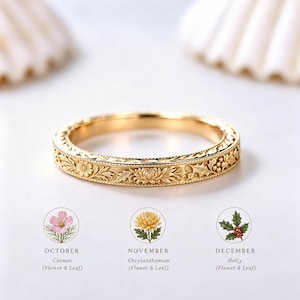 May include: A gold ring featuring an intricate engraved floral pattern. Below the ring, three circular icons display a pink cosmos for October, a yellow chrysanthemum for November, and a green holly sprig with red berries for December, each labeled.
