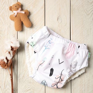May include: White baby diaper with a colorful feather and arrow pattern. A small brown teddy bear with a pink bow and a sprig of cotton are also in the image. The diaper has a soft, absorbent interior.