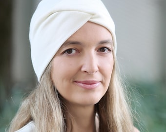 Puede incluir: Una diadema retorcida de color crema. La envoltura suave para la cabeza de tela la lleva una persona con cabello largo y castaño claro. La diadema es de color liso y parece estar hecha de un material suave y elástico. El fondo está borroso.