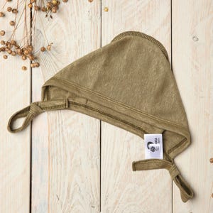 Puede incluir: Un gorro de bebé de punto verde con lazos. El gorro tiene una etiqueta que dice "ANA Handmade".