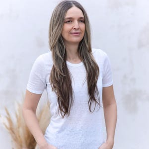 以下が含まれることがあります： 白い壁の前に立っている、長い茶色の髪の女性が白いTシャツを着ています。