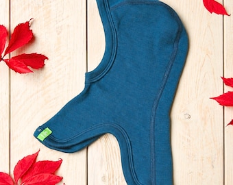 May include: A deep blue balaclava hood made of a soft knit fabric with visible seam detailing along the crown and neck. The garment features a small, bright green rectangular tag attached to the lower edge, providing a distinct color contrast.