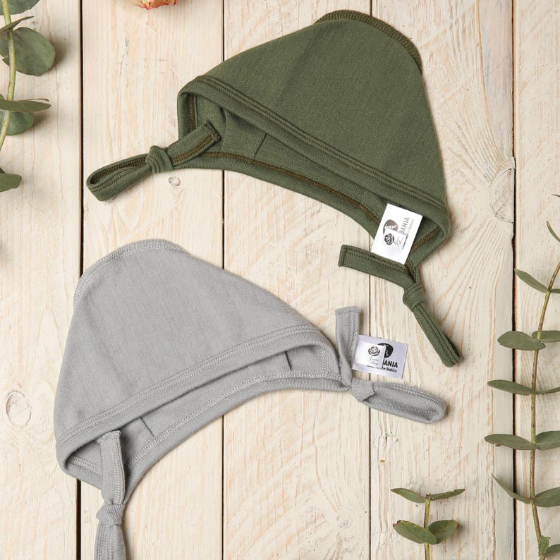May include: Two baby bonnets, one in a light gray color and one in a dark green color. Both bonnets have a tie at the back and a small label with the word "BANIA" on it.