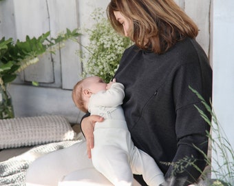May include: A woman wearing a dark grey sweatshirt is holding a baby in a white onesie. The baby is looking at the woman and the woman is looking down at the baby. The woman is sitting on a white and grey blanket.