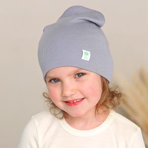 May include: A light gray beanie hat made of a smooth, soft-knit fabric. The beanie features a small, square white label on the front right side with a green rose icon and the text "Green Rose" printed in a clear, dark green font.