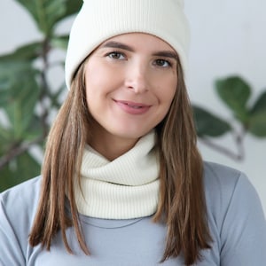Puede incluir: Una mujer con un gorro de punto blanco y un cuello de punto blanco. Está sonriendo y mirando a la cámara.