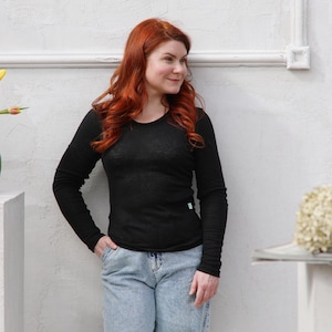 Puede incluir: Una camiseta negra de manga larga con cuello redondo. La camiseta se combina con vaqueros azul claro. La modelo está de pie frente a una pared blanca.