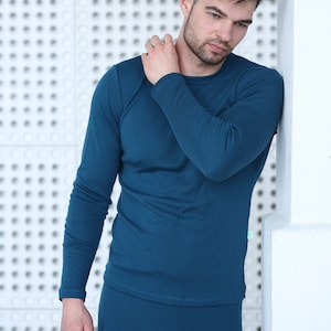 May include: Teal long-sleeved top and matching trousers. The top has a crew neck and the trousers are form-fitting. The outfit is made of a soft, stretchy material. The person is leaning against a white wall with a textured pattern.