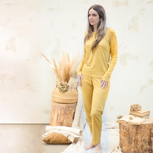 Op de afbeelding: Een vrouw draagt een mosterdgele, langarmige top en een bijpassende broek. De top heeft een ronde hals en de broek heeft een rechte pijp.