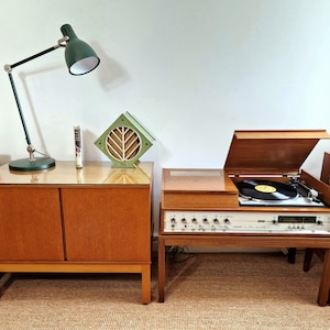 Könnte beinhalten: Ein Vintage-Entertainment-Center mit einem Plattenspieler, Lautsprechern und einem Schrank. Eine grüne Schreibtischlampe und ein dekoratives grünes und hölzernes Akzentstück stehen auf dem Schrank. Das Set steht auf einem braunen Teppich.