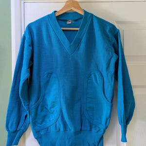 Puede incluir: Una sudadera vintage de color azul verdoso con cuello en V y mangas largas. La sudadera tiene dos bolsillos grandes y redondeados en la parte delantera y puños y dobladillo acanalados. La prenda está colgada de una percha de madera contra una puerta blanca.