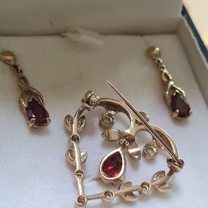 May include: A gold heart-shaped brooch with red gemstone accents and a delicate leaf design. The brooch is accompanied by a pair of matching earrings.