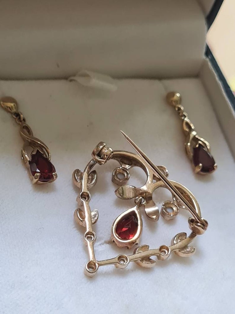May include: A gold heart-shaped brooch with red gemstone accents and a delicate leaf design. The brooch is accompanied by a pair of matching earrings.
