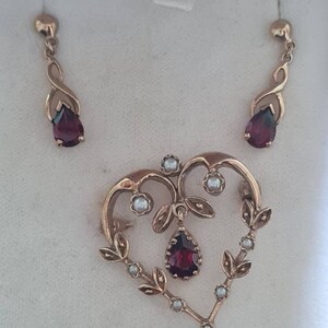 May include: A heart-shaped pendant with a dangling red gemstone and pearl accents. The pendant is part of a set with matching earrings featuring red gemstones.