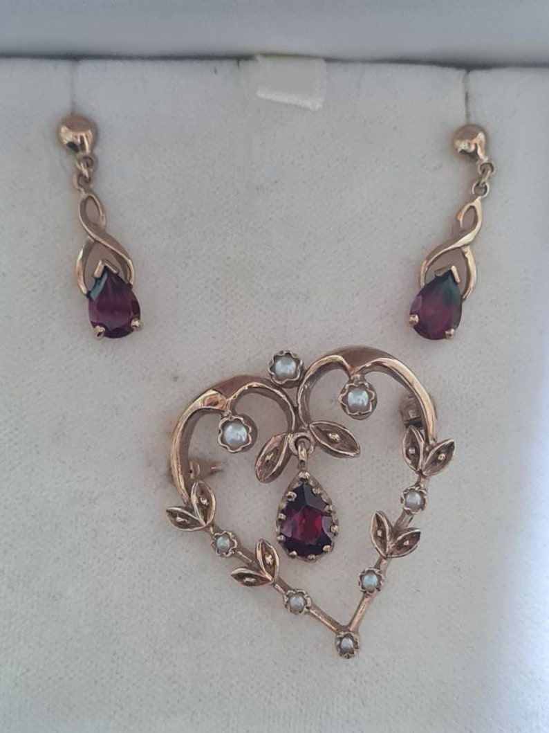May include: A heart-shaped pendant with a dangling red gemstone and pearl accents. The pendant is part of a set with matching earrings featuring red gemstones.