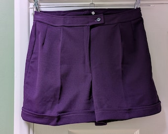 May include: A pair of purple shorts with a button closure and pleats. The shorts are hanging on a silver hanger against a white door. The shorts have a cuffed hem and a tailored design.