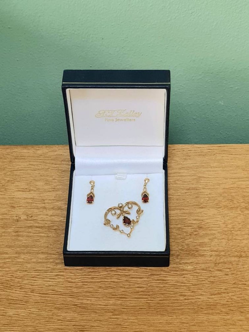 May include: A heart-shaped pendant and matching earrings with red gemstones and small white pearls. The jewelry is displayed in a black box with the text "S.J. Bailey Fine Jewellers".