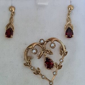 May include: A gold heart-shaped pendant with a red gemstone and small white pearls. The pendant is part of a set with matching earrings.