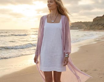 May include: A woman wearing a white linen dress and a long, light pink cardigan on a beach. She is also wearing a silver necklace and bracelet. The background features the ocean and a sunset.
