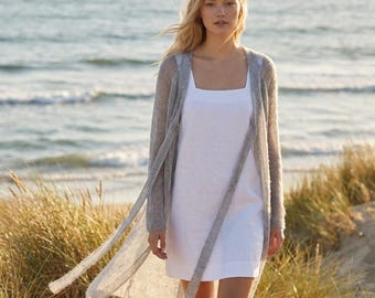 May include: A woman wearing a white square-neck dress and a long, light gray cardigan with long ties. The outfit is worn on a sandy beach with the ocean in the background. The cardigan is a lightweight knit.