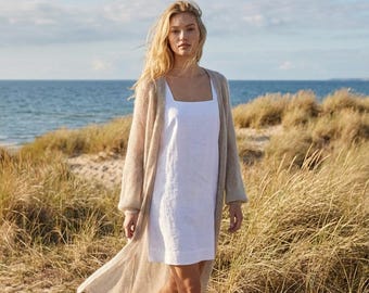 May include: A woman wearing a white linen dress and a long, beige cardigan. The dress has a square neckline. The cardigan is open and flowing. The background features a beach with tall grass and the ocean.