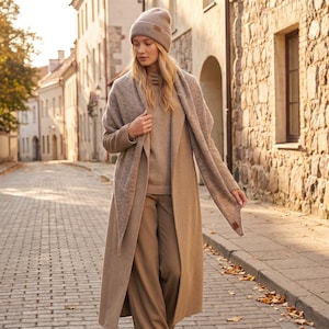 May include: A beige knit beanie, a matching beige knit scarf, a beige turtleneck sweater, and a long beige coat. The beanie and scarf feature a small rectangular brown leather-like patch. The outfit is completed with tan trousers.