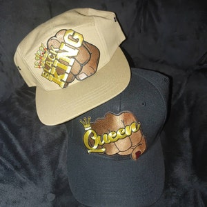 Peut inclure: Deux casquettes de baseball, l'une beige et l'autre noire, avec des motifs de poing brodés. La casquette beige a une couronne dorée et les mots "Black King" sur le poing. La casquette noire a une couronne dorée et le mot "Queen" sur le poing.