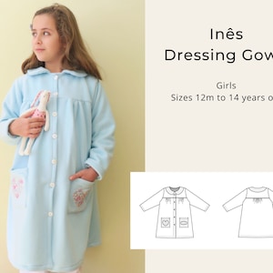 Op de afbeelding: Een lichtblauwe badjas met een witte kraag, knopen en zakken. De badjas heeft een bloemenpatroon op de zakken. De tekst "Inés Dressing Gown" en "Girls Sizes 12m to 14 years old" is zichtbaar in de afbeelding.