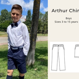 Op de afbeelding: Een jonge jongen draagt een wit overhemd met knopen en een donkerblauwe strik, en donkerblauwe shorts. De jongen staat in een park met groen gras en bomen. De afbeelding bevat ook een lijntekening van een broek en een short. De tekst "Arthur Chinos" en "Boys" is zichtbaar in de afbeelding. De tekst "Sizes 3 to 15 years old" is ook zichtbaar.