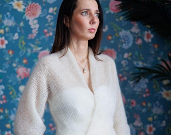 May include: A woman wearing a white, open-front, short-sleeved sweater over a white dress. The sweater has a soft, textured appearance. The background features a floral wallpaper and a pink sofa.