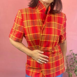 May include: A short-sleeved blazer featuring a vibrant red, yellow, and orange plaid pattern. The garment has a high collar, a button-front closure, and two horizontal pockets on the lower front section, constructed with a structured fit.