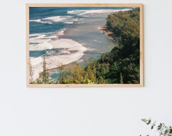 May include: A framed photograph of a beach scene with turquoise water crashing on the shore. Lush green trees and foliage line the coastline, creating a vibrant contrast against the blue water.
