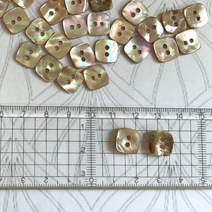 Könnte beinhalten: Eine Sammlung quadratischer, schillernder Knöpfe mit zwei Löchern. Die Knöpfe sind hellbraun und cremefarben mit Perlmuttglanz. Die Knöpfe liegen auf einem karierten Hintergrund mit Zentimetermessungen.