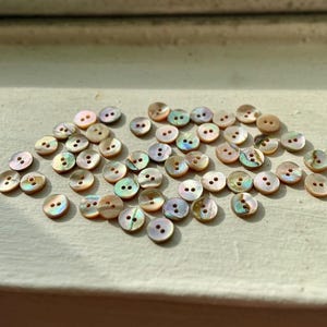 May include: A collection of small, round buttons with iridescent, pearlescent surfaces. Each button has two holes for sewing. The buttons vary in color, with shades of pink, green, and gold. They are scattered on a light-colored surface.