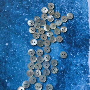 May include: A selection of round, iridescent buttons with two or four holes, arranged on a blue backdrop. The buttons have a pearlescent sheen, reflecting light. These buttons are suitable for sewing and craft projects.